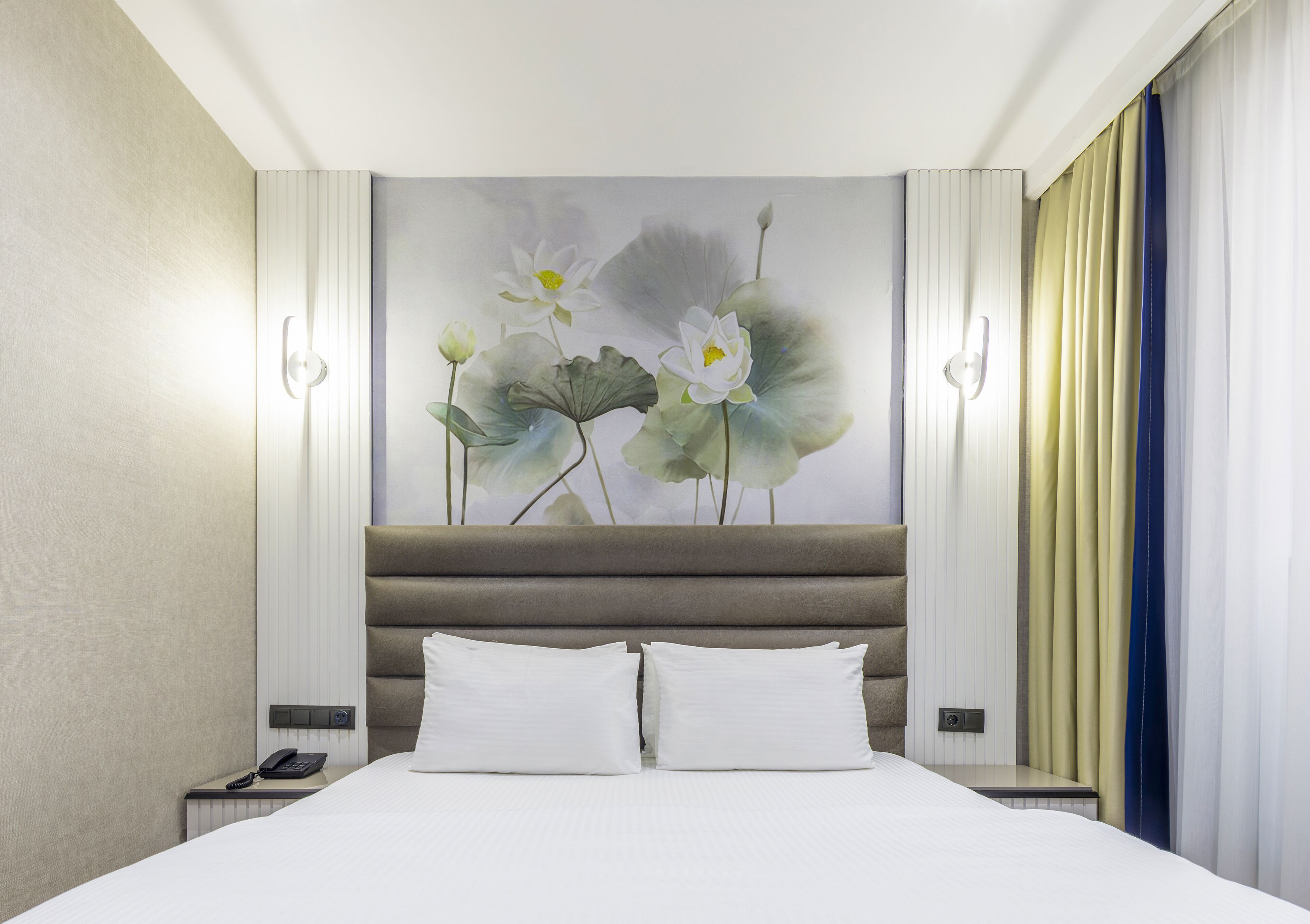 A hotel room with a large bed, two bedside tables, a nightstand, a painting of flowers, and a view of the city.