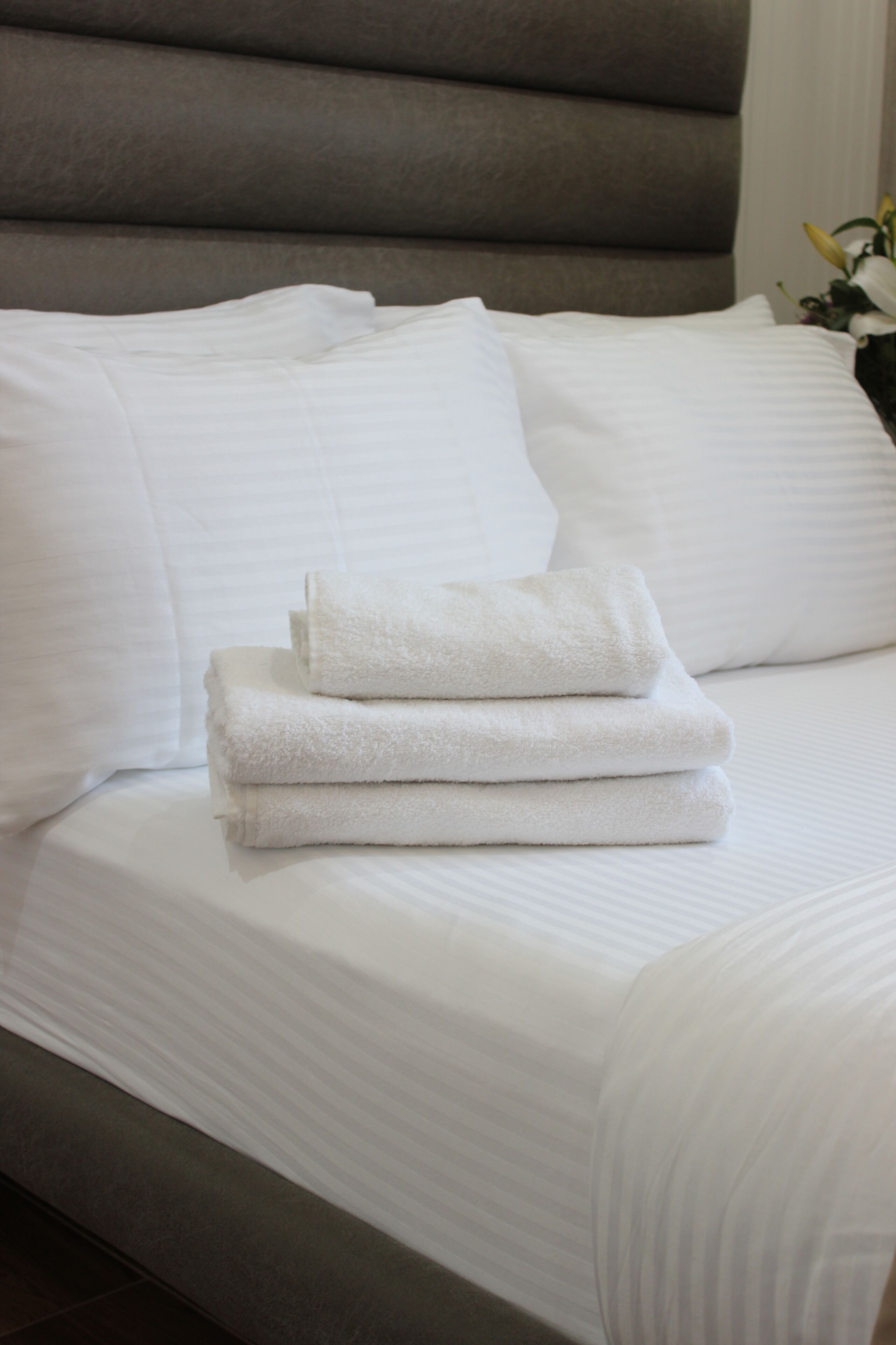 A neatly made bed with white linens and folded towels, a large headboard, and a vase with flowers on the side.