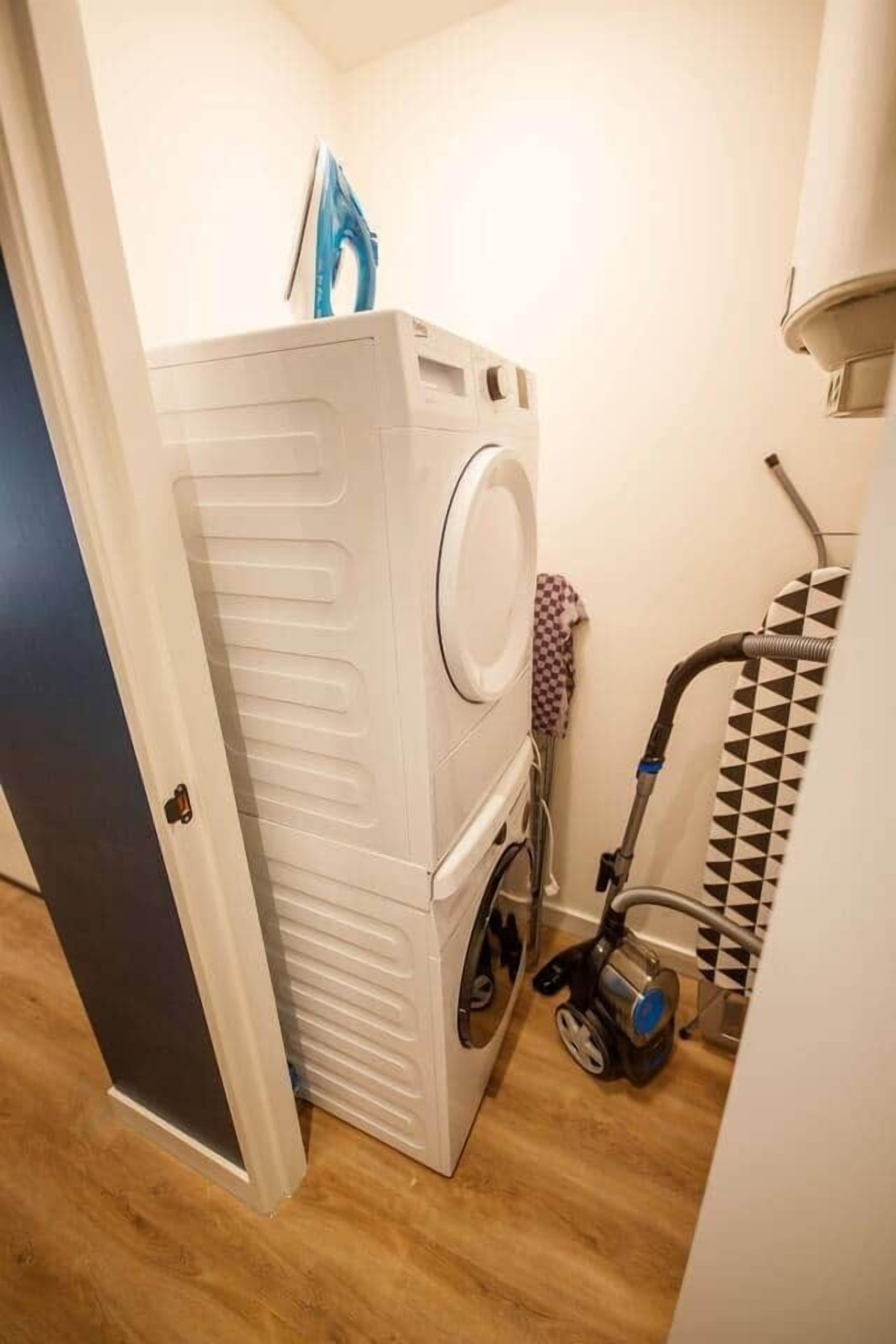 A compact laundry area with a stacked washer and dryer, a vacuum cleaner, and a patterned towel rack.
