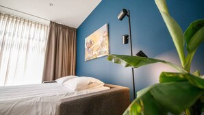A hotel room with a bed, a painting on the wall, a floor lamp, and a large plant.