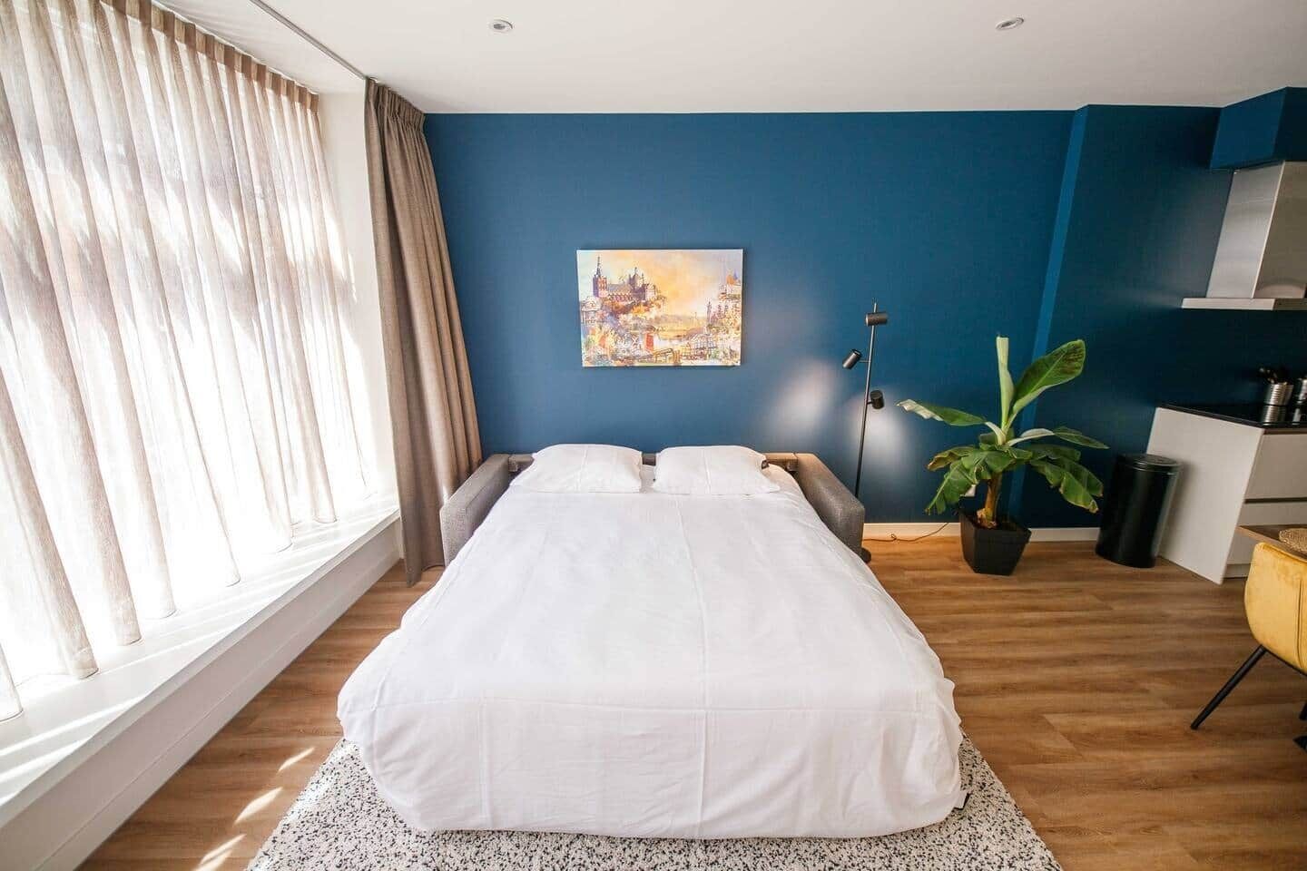 A modern bedroom with a bed, a cityscape painting, a potted plant, and a desk with a chair.