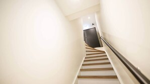 Una escalera estrecha y bien iluminada con escalones de madera y barandilla, que lleva a una puerta oscura al final.