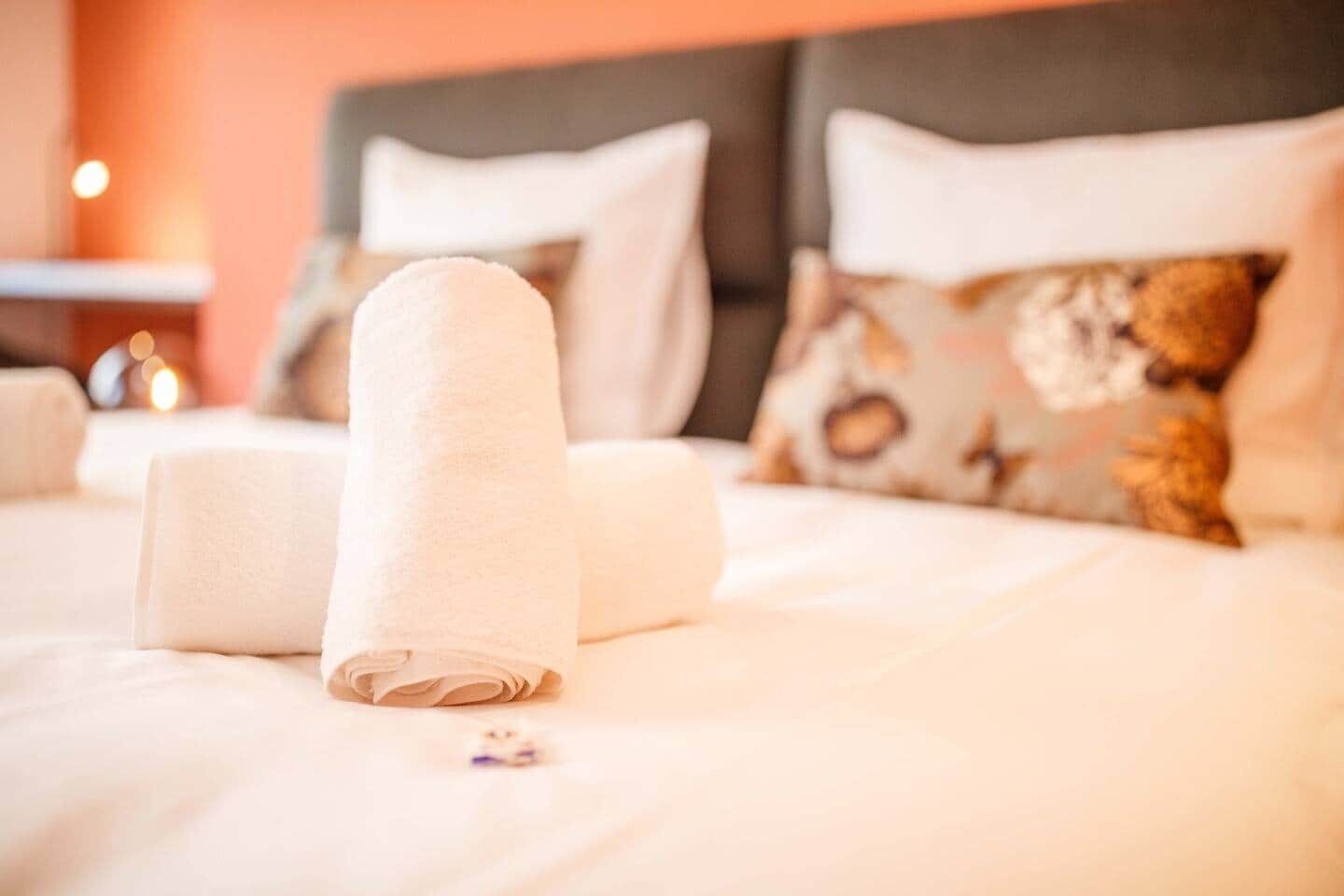 Ein ordentlich bezogenes Bett mit weißen Leinentüchern und einem gerollten Handtuch, ein Kissen mit Bärenmotiv und ein Nachttisch mit einer Lampe.