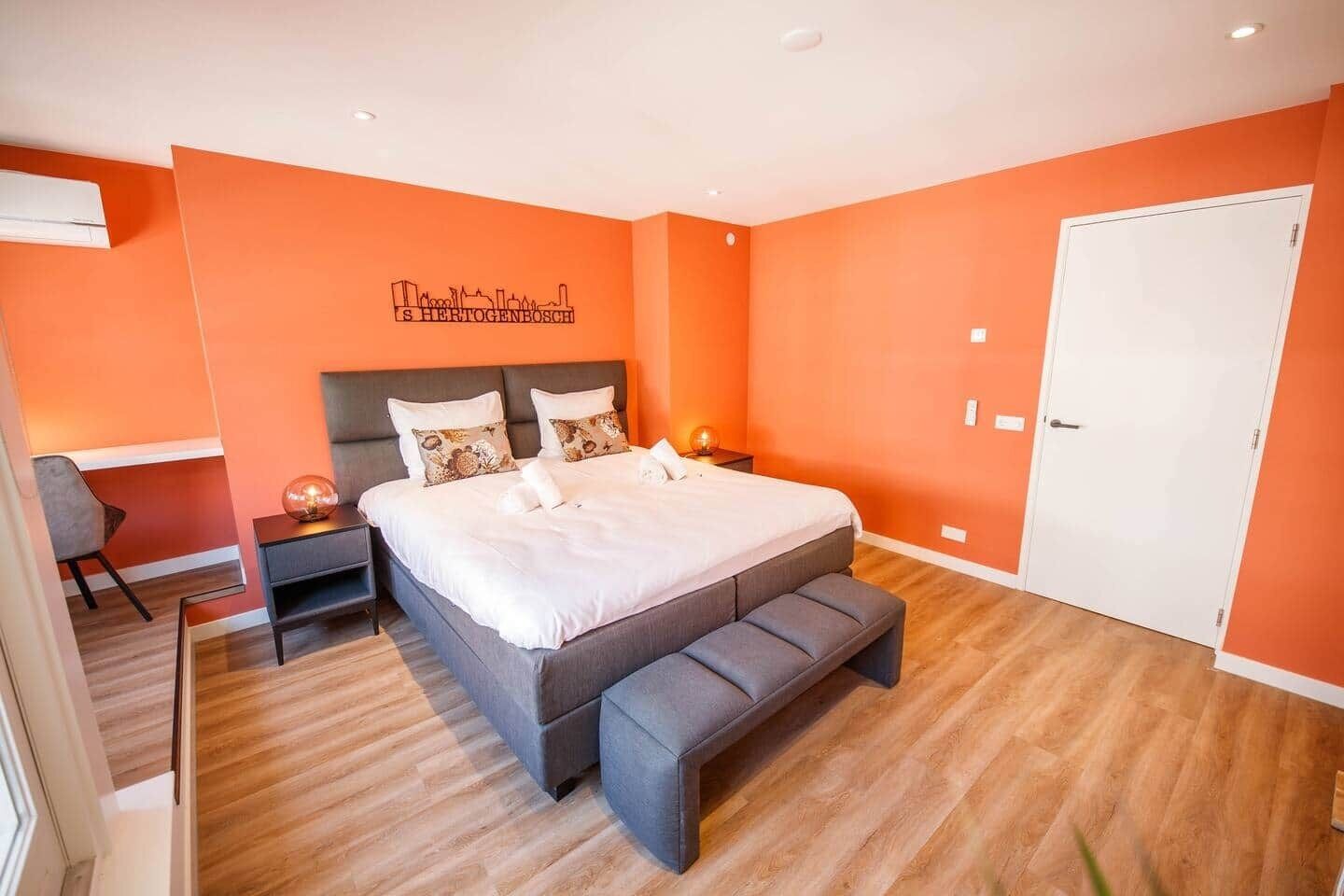 Une chambre moderne avec un grand lit, un banc, un bureau et une fresque représentant un paysage urbain sur le mur.