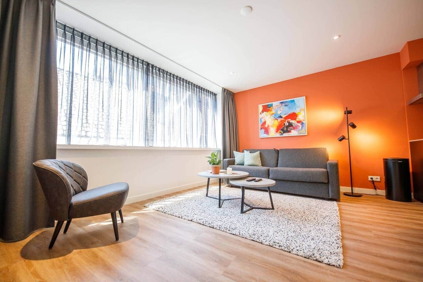Una sala de estar moderna con un sofá gris, un sillón marrón, una mesa de centro blanca y un ventanal con cortinas translúcidas.