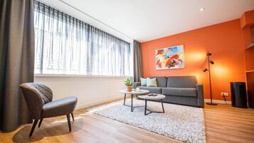Una sala de estar moderna con un sofá gris, un sillón marrón, una mesa de centro blanca y un ventanal con cortinas translúcidas.
