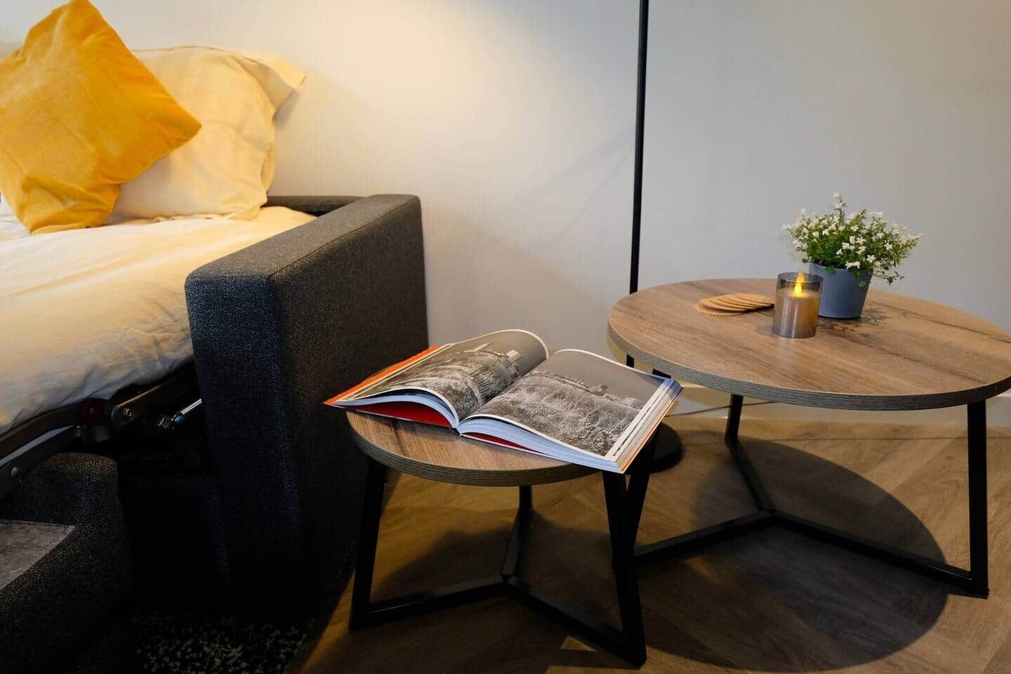 A modern hotel room with a bed, a reading chair, a small round table with a candle, and a potted plant.