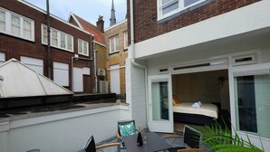 A small outdoor patio with a table and chairs, a bed with a window, and a brick building in the background. - Luxury 60m2 Apartment with Terrace (R-65-A) (Eindhoven)