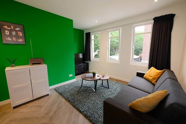 A modern living room with a green accent wall, a grey sofa with yellow cushions, a small round table, a cabinet, and a television. - Luxury 60m2 Apartment with Terrace (R-65-A) (Eindhoven)