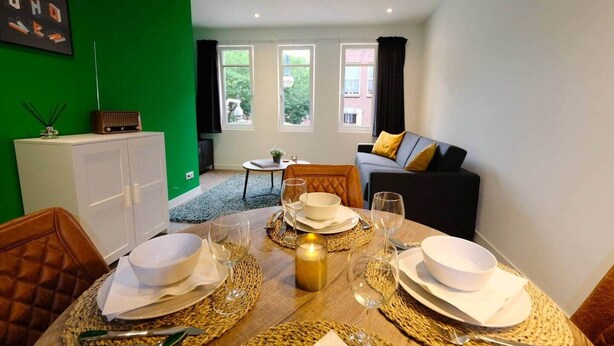 A dining area with a table set for four, a sofa, and a cabinet in a room with green walls and large windows. - Luxury 60m2 Apartment with Terrace (R-65-A) (Eindhoven)