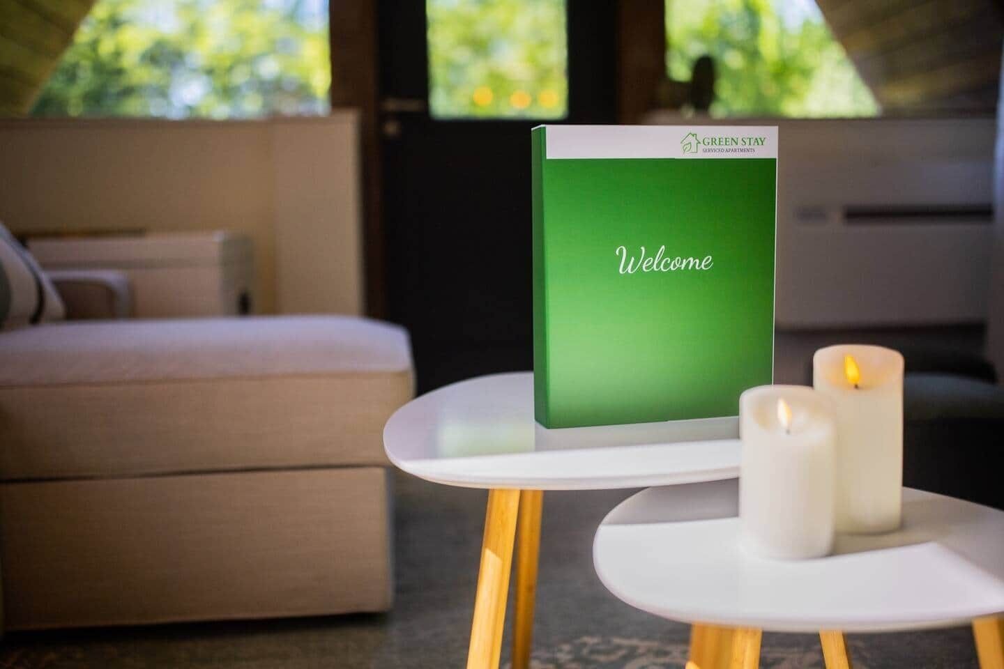 Un panneau vert avec le mot « Bienvenue » est posé sur une table blanche dans une pièce où se trouvent un canapé et deux bougies allumées.