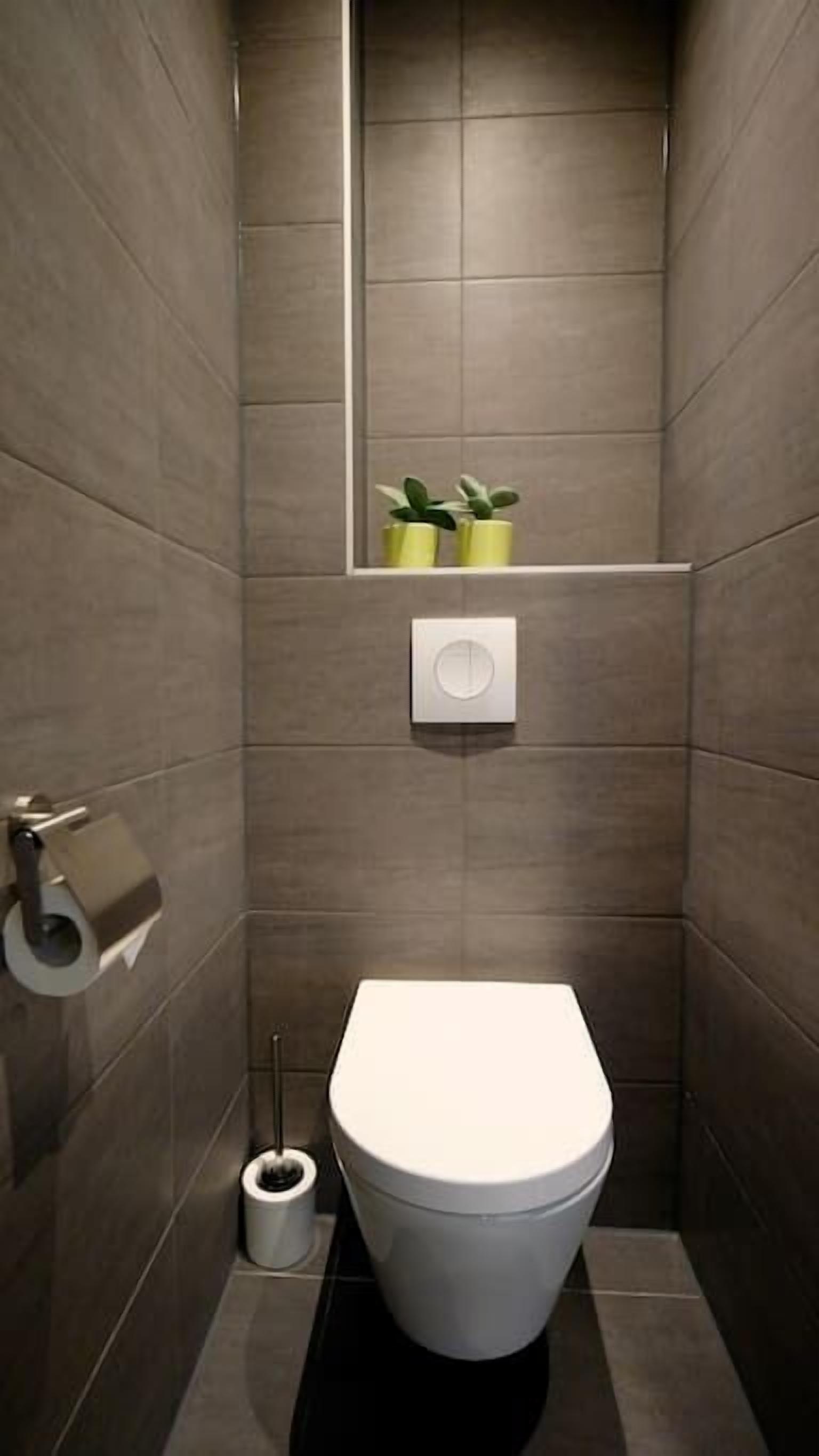 Un baño moderno con inodoro, dispensador de papel higiénico, un pequeño cubo de basura y un espejo con una planta.