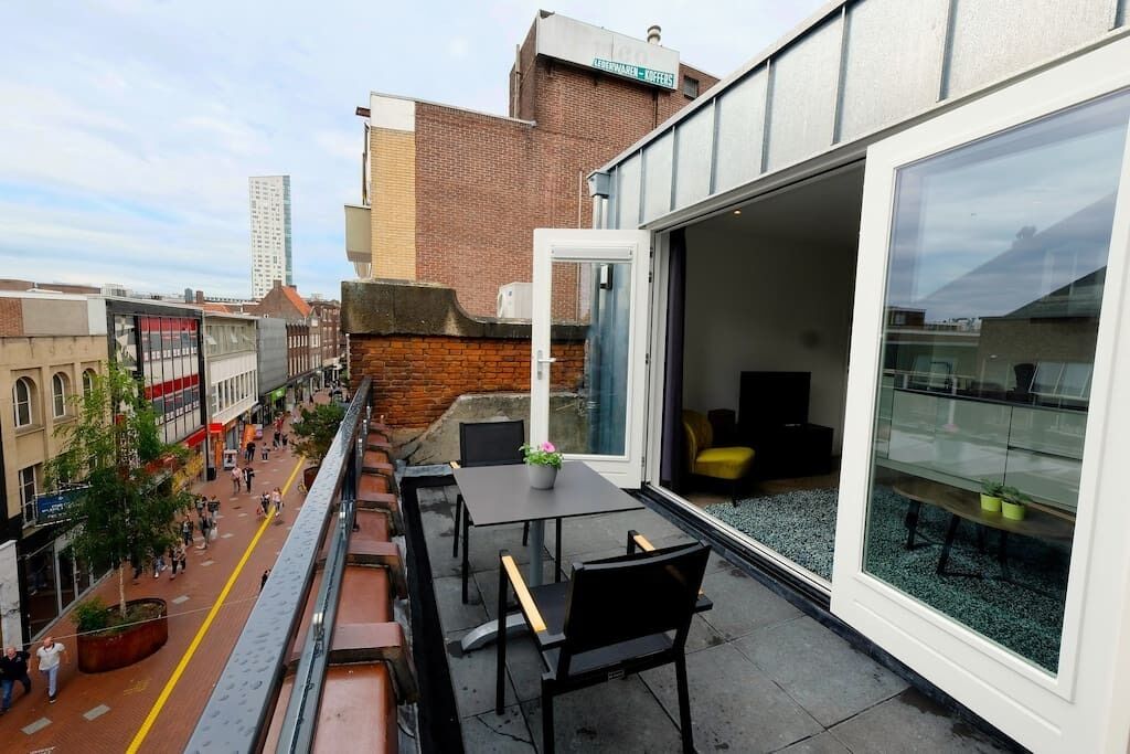 An outdoor seating area with tables and chairs, a glass door leading to an interior space, and a view of a city street with buildings and pedestrians.
