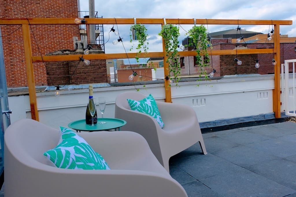 A rooftop terrace with two white chairs, a small table with a bottle and glass, and a wooden pergola with hanging lights.