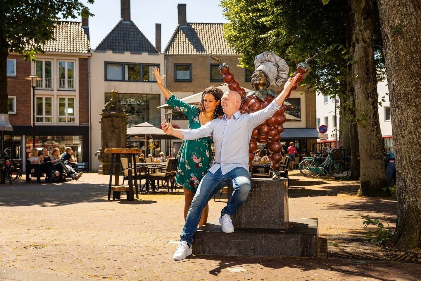 Un couple pose devant une sculpture sur une place publique, avec des bâtiments en arrière-plan.