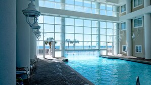 Une grande piscine intérieure avec vue sur l’océan, entourée d’un espace couvert aménagé avec des sièges et un plafond en verre.