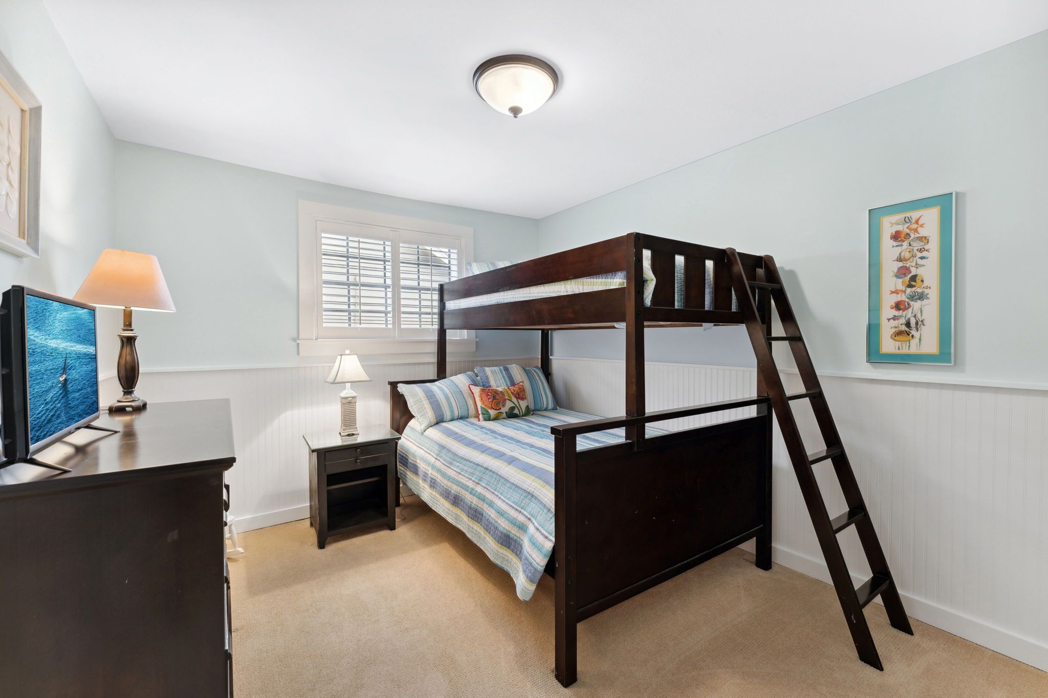 A bunk bed with a ladder, a desk with a lamp, a TV, and a window with blinds.