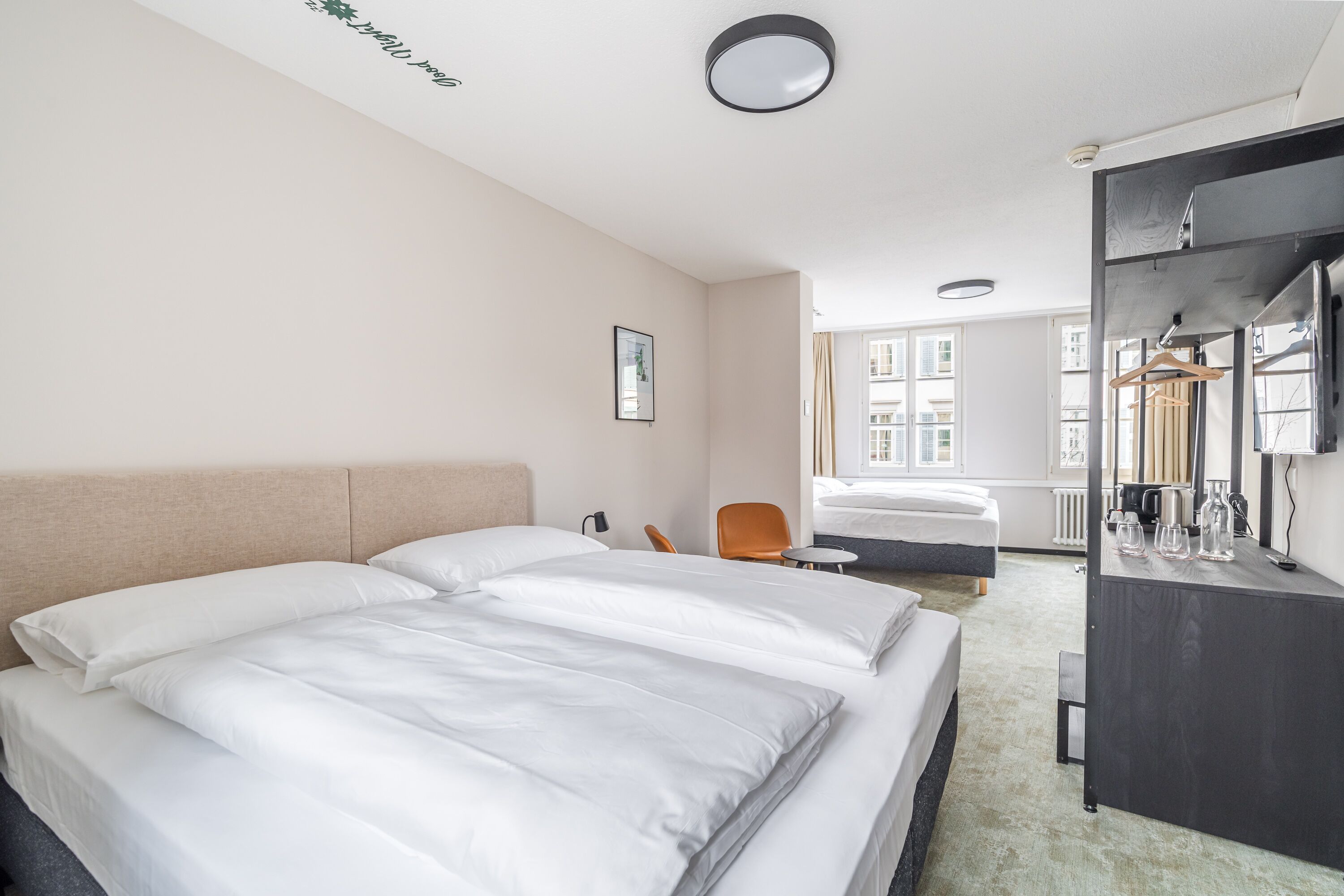 Ein Hotelzimmer mit zwei Betten, einem Schreibtisch, einem Stuhl, einem Spiegel und einem Fenster mit Blick auf Gebäude.