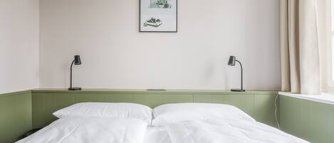 Een net opgemaakt bed met witte lakens, een ingelijst schilderij aan de muur en twee bedlampjes.
