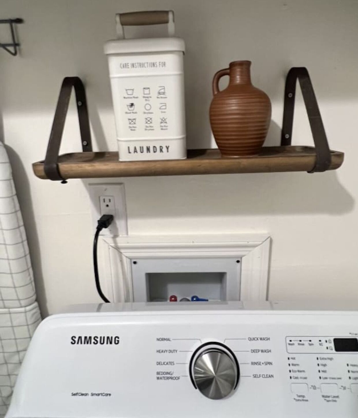 A Samsung self-cleaning smart core washing machine with a control panel, a care instructions box, and a brown vase on a shelf above.