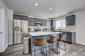 A modern kitchen with grey cabinets, stainless steel appliances, a large island with bar stools, and a window providing natural light. - Countryside Classic with Firepit (Nampa)
