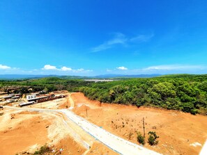 Una obra en construcción con camino de tierra, edificios y zona boscosa bajo un cielo azul despejado.