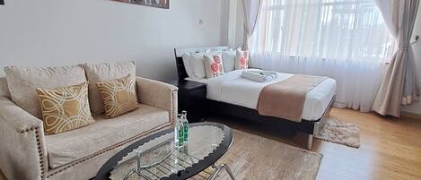 A modern hotel room with a sofa, a bed with a white and brown bedspread, a glass coffee table, and a large window with sheer curtains.
