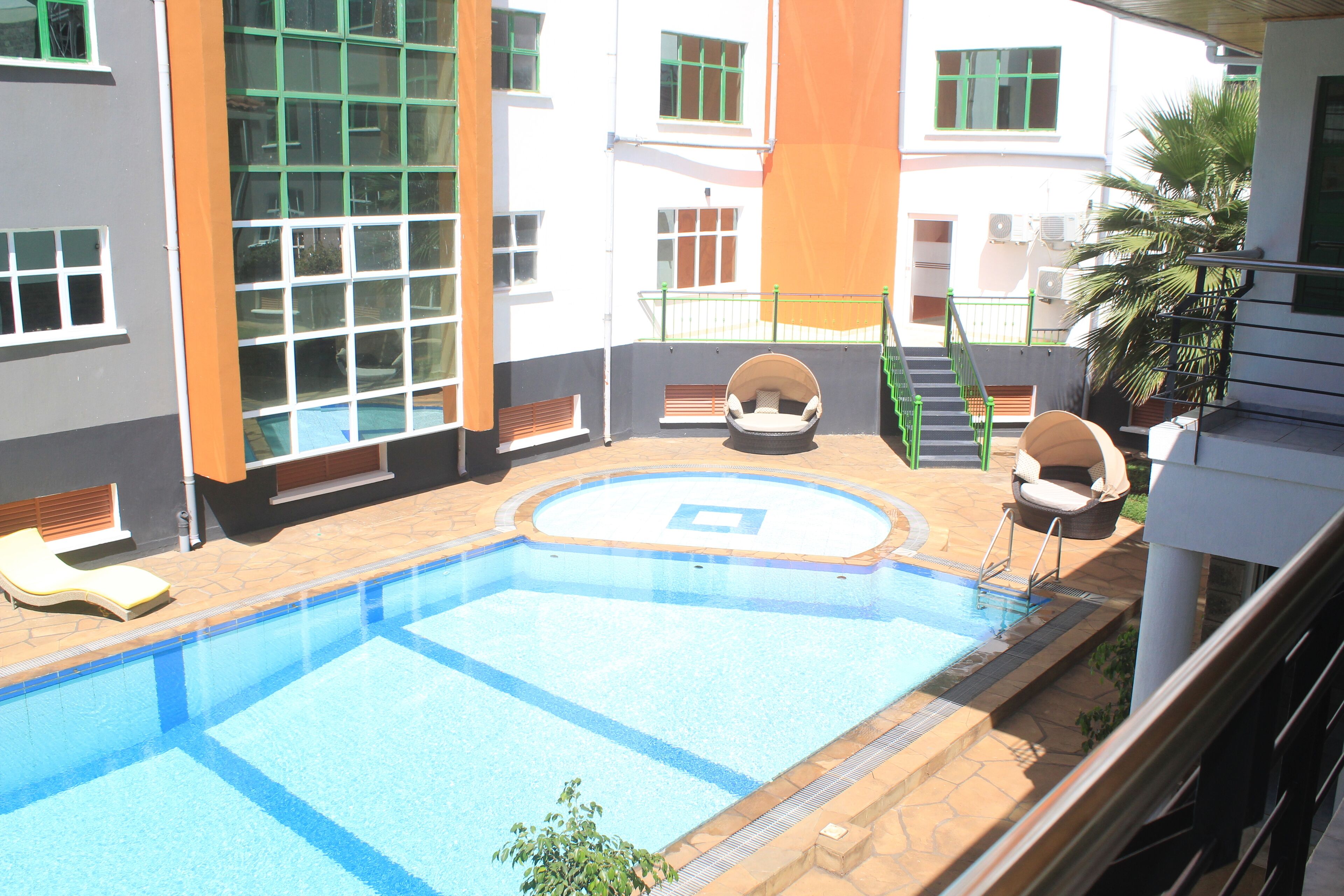 A swimming pool with a sunbathing area, a staircase, and a view of a building with large windows and a palm tree.