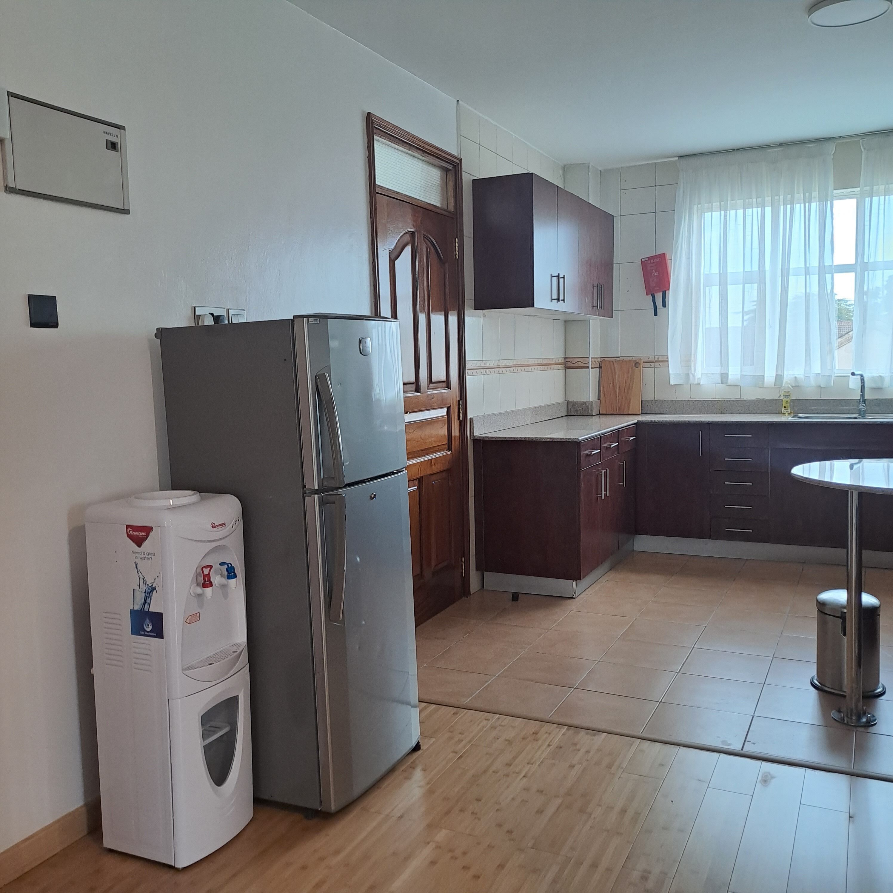 Een keuken met een roestvrijstalen koelkast, een waterdispenser, houten kastjes, een spoelbak en een raam met transparante gordijnen.