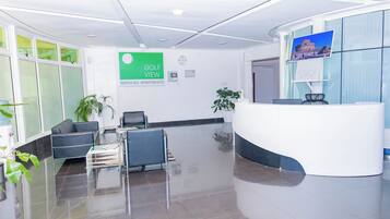 Una moderna zona de recepción con un mostrador blanco curvo, un área de descanso con sillas y un amplio ventanal con cortinas verdes.