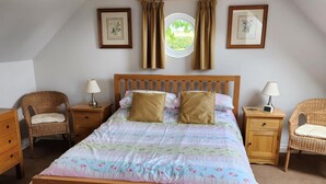Een slaapkamer met een houten bed, twee nachtkastjes, twee rieten stoelen, een raam met gordijnen en ingelijste foto’s aan de muur.