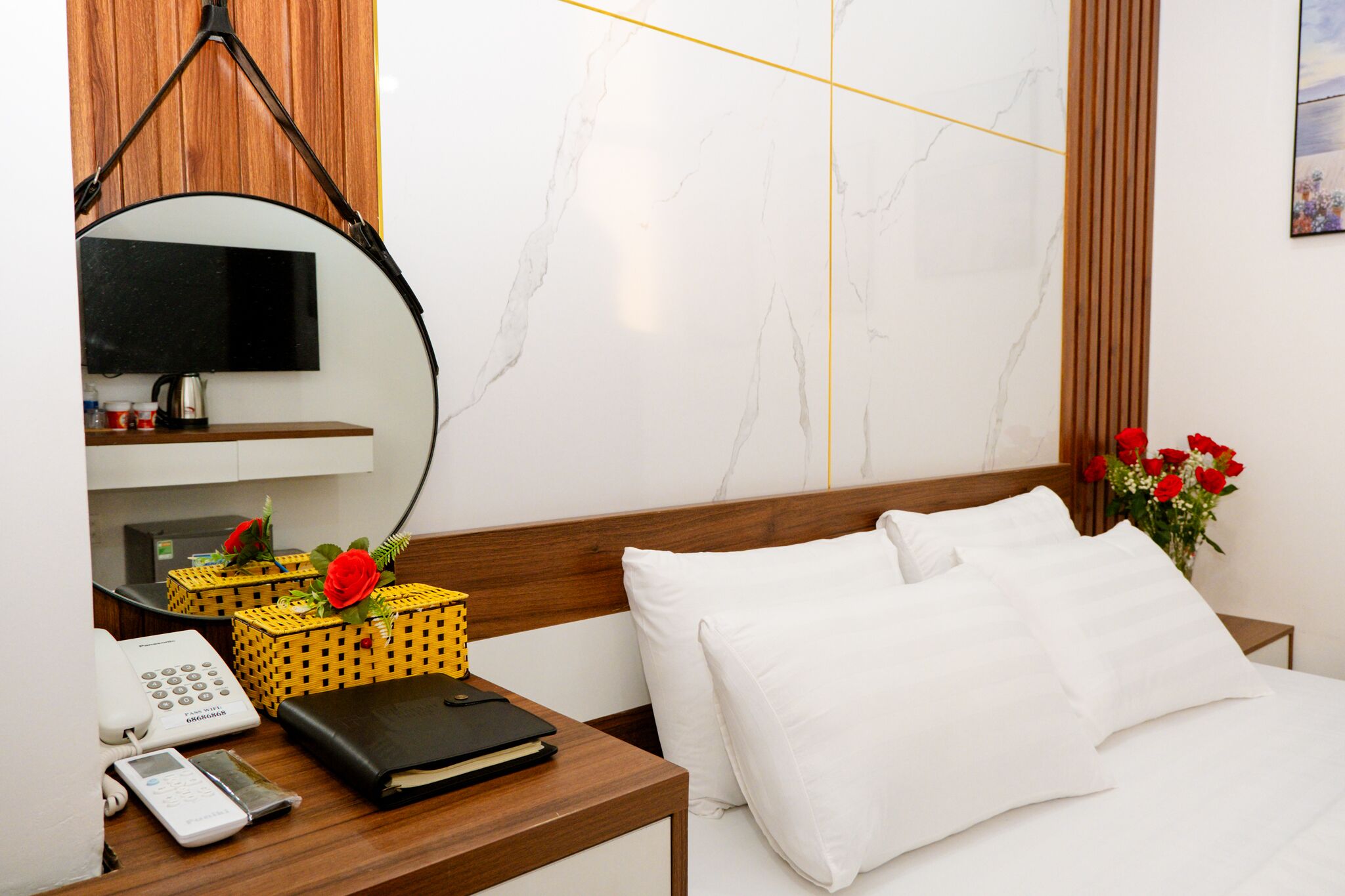 Une chambre d’hôtel avec une tête de lit en bois, un lit recouvert de draps blancs, une table de chevet avec un téléphone, une horloge numérique, un carnet noir et un panier jaune contenant des fleurs rouges.