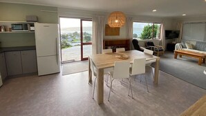 A modern kitchen with a dining area, large windows, and a view of the outdoors. - Little Aotea bach with sea views (Kawhia)