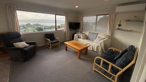 A spacious living room with a large window, a flat-screen TV, a wooden coffee table, and two sofas. - Little Aotea bach with sea views (Kawhia)