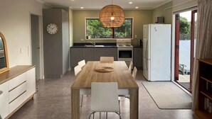 A modern kitchen with a dining area, a clock, and a view of greenery outside. - Little Aotea bach with sea views (Kawhia)
