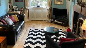 Ett vardagsrum med en svart soffa, en svart fåtölj, ett soffbord i glas, en svartvit matta med sicksackmönster, en tv, en öppen spis och ett stort fönster med gardiner.
