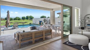 Ein modernes Wohnzimmer mit einem großen Schiebeglastür, die zu einem Poolbereich führt, einer Holzbank und einem Esstisch mit Stühlen.