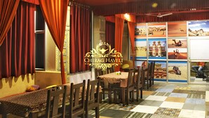 A restaurant interior with red curtains, wooden chairs, a tiled floor, and a ceiling fan. - Chirag Haveli - Near Golden Fort (Jaisalmer)