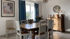 Comedor con una mesa de madera, sillas blancas, un florero con flores, un reloj y una obra de arte enmarcada en la pared.