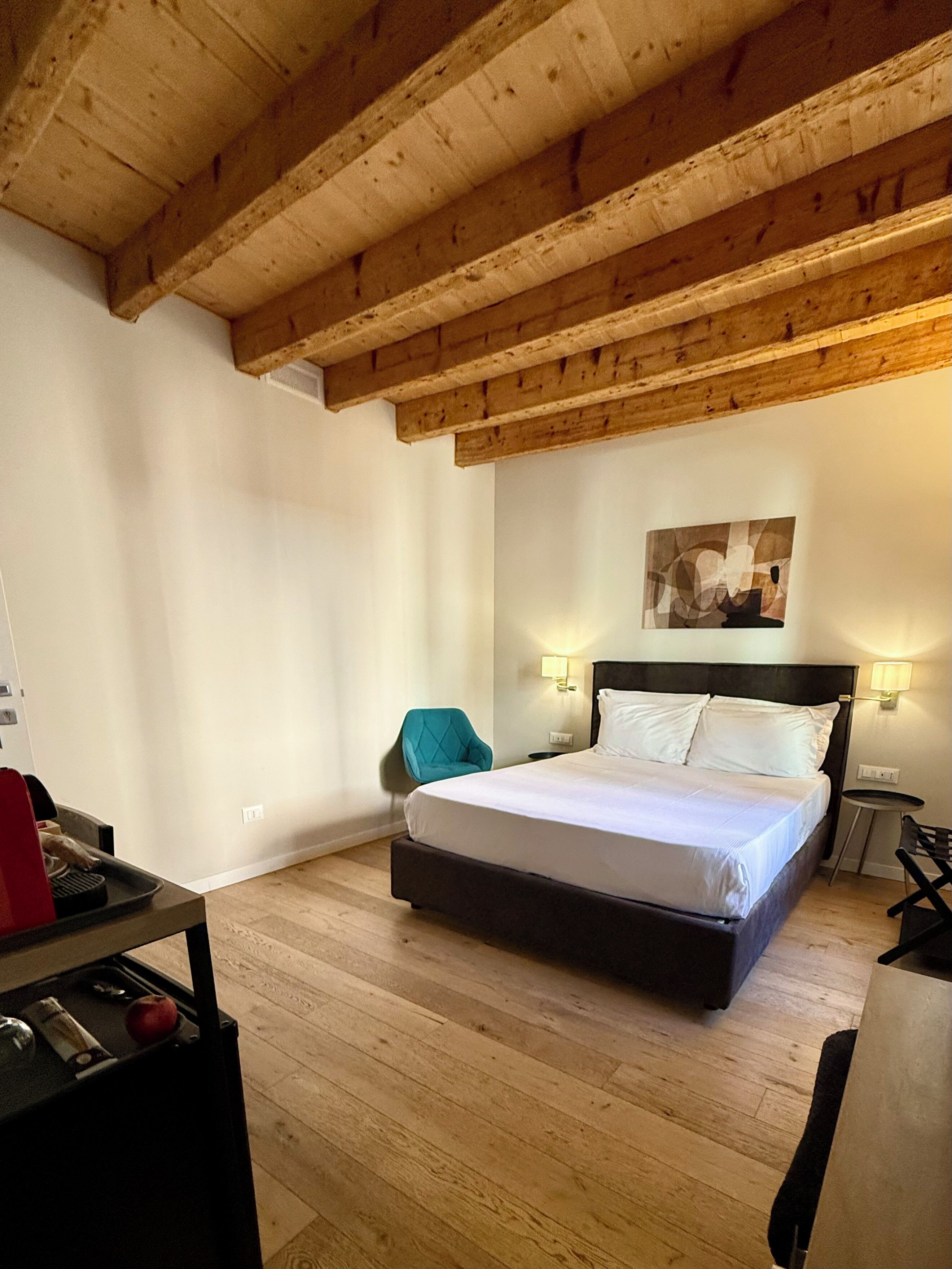 A bedroom with a wooden ceiling, a bed with white linens, a teal chair, and a small table with a red object.