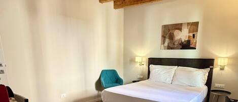 A bedroom with a wooden ceiling, a bed with white linens, a teal chair, and a small table with a red object.