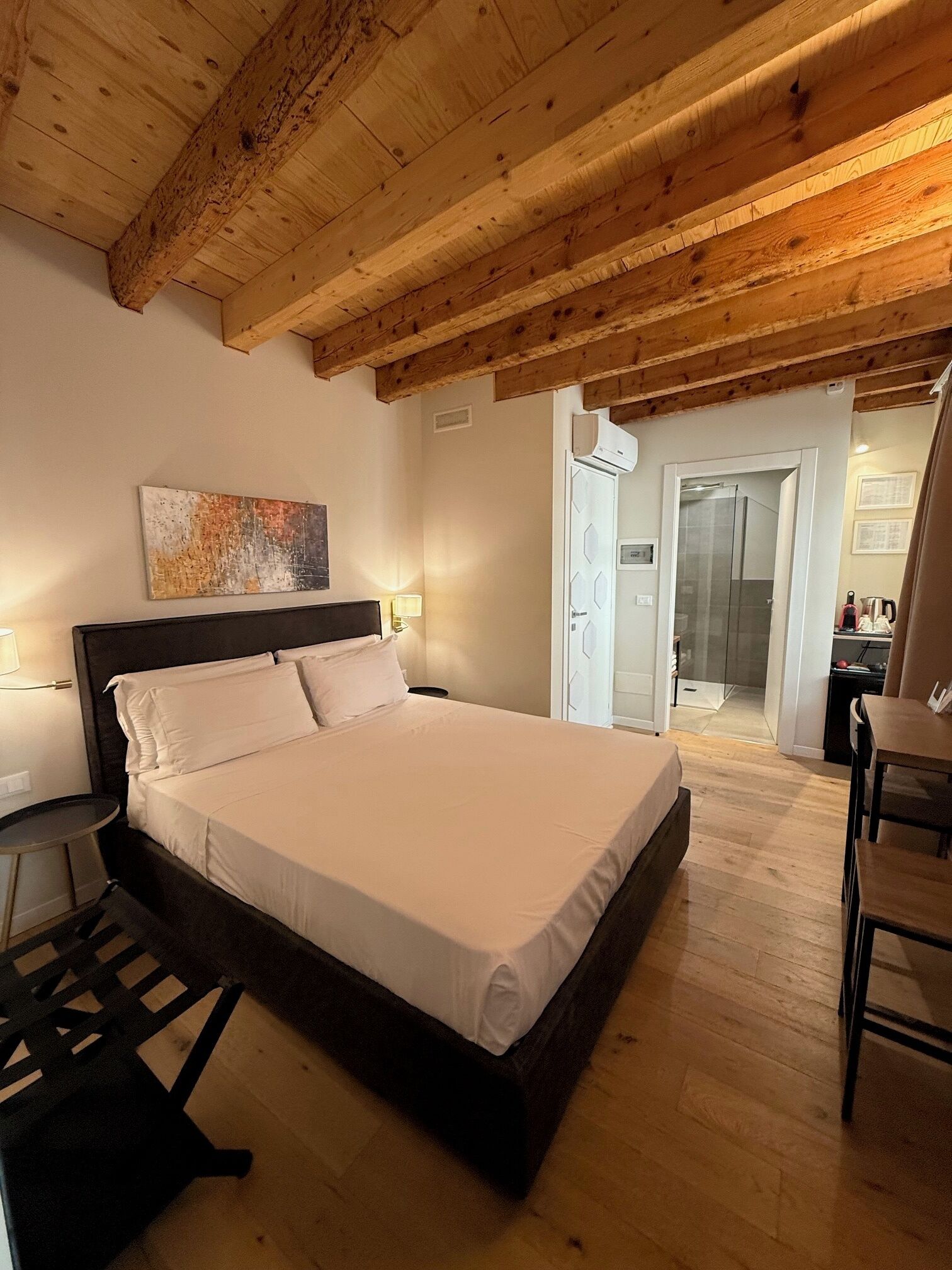 Ein Schlafzimmer mit Holzbalkendecke, einem Bett, einem Nachttisch, einem Sessel und einem durchgehenden Türspalt erkennbaren Badezimmer.