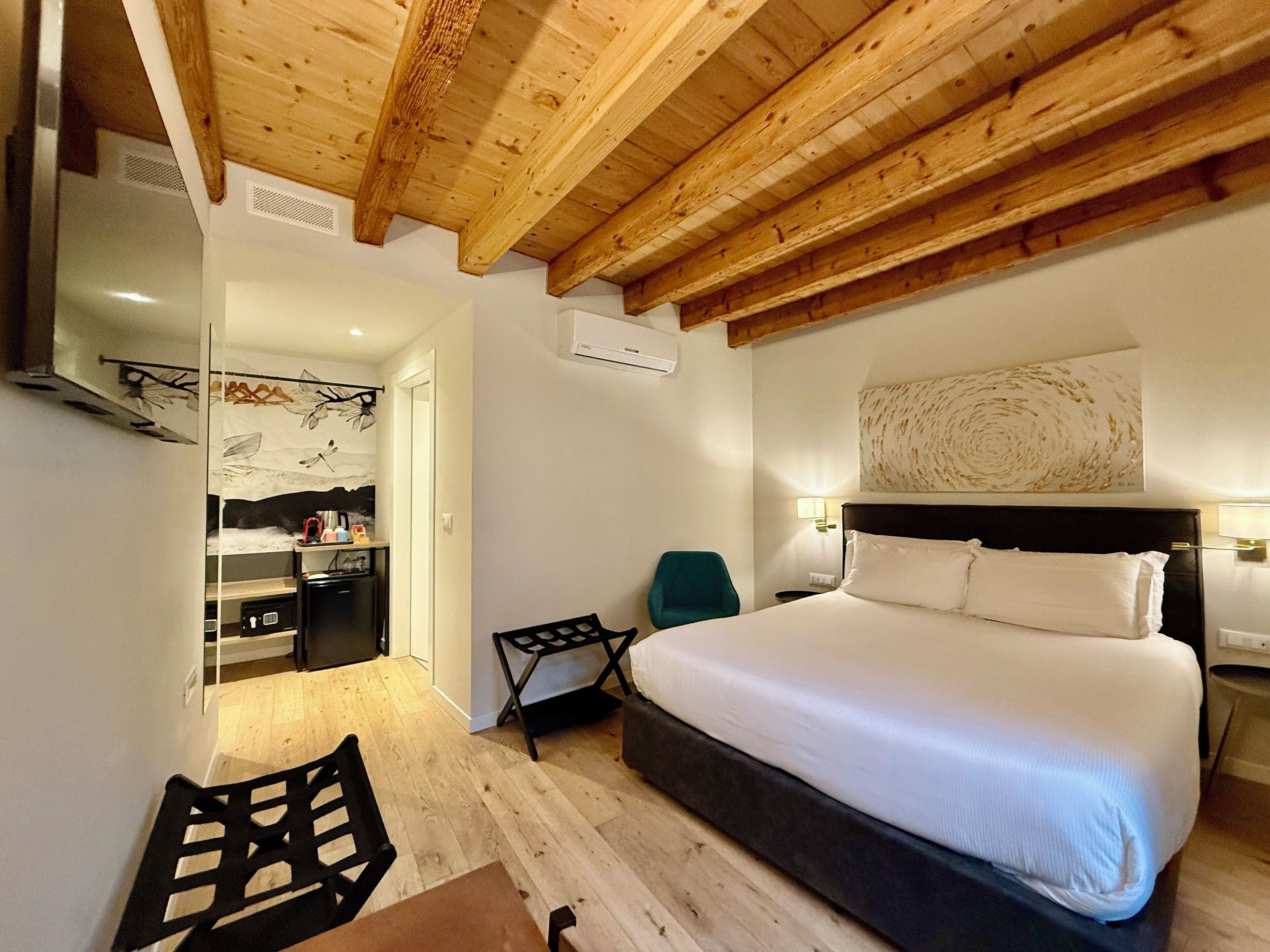 Habitación de hotel con techo de madera, cama con ropa blanca, escritorio con silla y una kitchenette pequeña.
