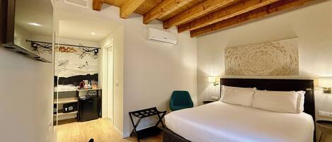 A hotel room with a wooden ceiling, a bed with white linens, a desk with a chair, and a small kitchenette.
