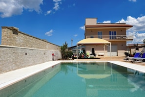 Une piscine bordée d’un mur en pierre, un espace de repos couvert et un bâtiment doté de balcons.