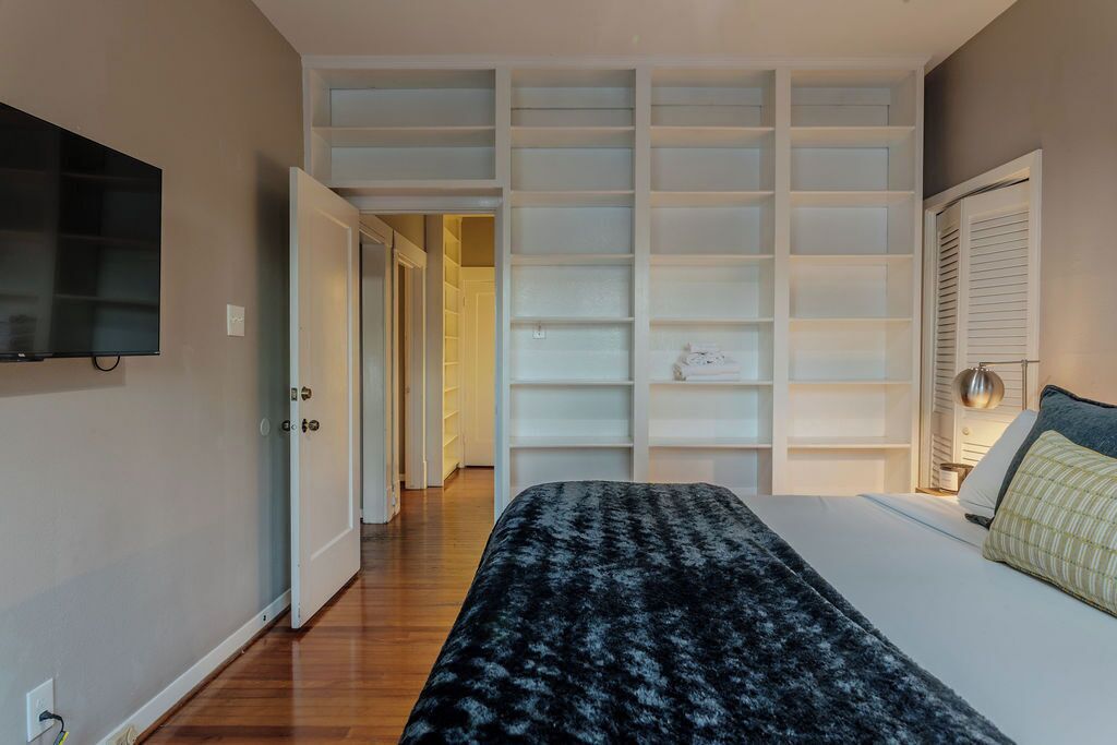 Ein Schlafzimmer mit Bett, einem an der Wand befestigten Fernseher, einem begehbaren Kleiderschrank mit Regalen und einer Tür zu einem anderen Raum.