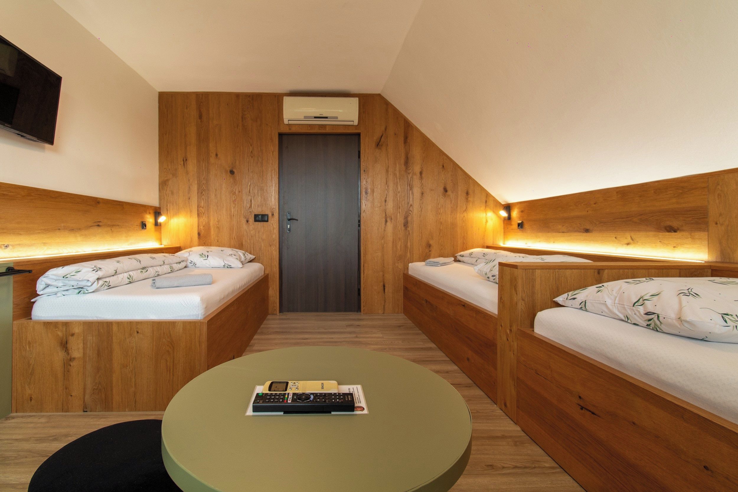 A compact hotel room with two wooden beds, a small table, and a television mounted on the wall.