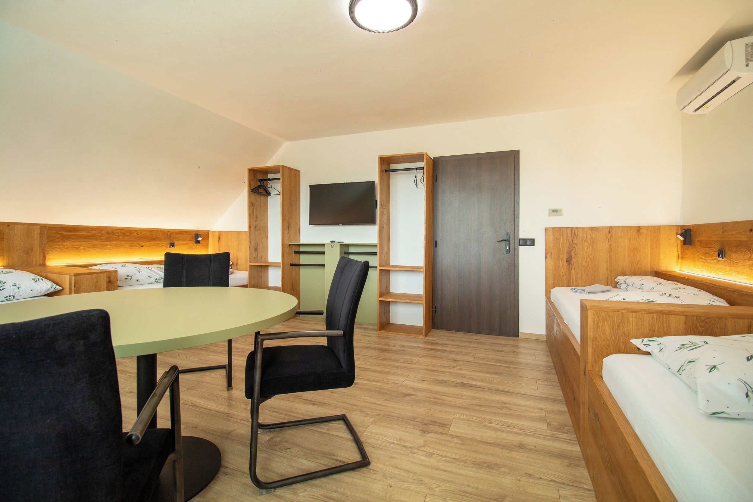 Una habitación de hotel con piso de madera, una mesa redonda con sillas, una cama con cabecera, un televisor montado en la pared y una puerta que da a otra habitación.