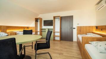 Una habitación de hotel con piso de madera, una mesa redonda con sillas, una cama con cabecera, un televisor montado en la pared y una puerta que da a otra habitación.
