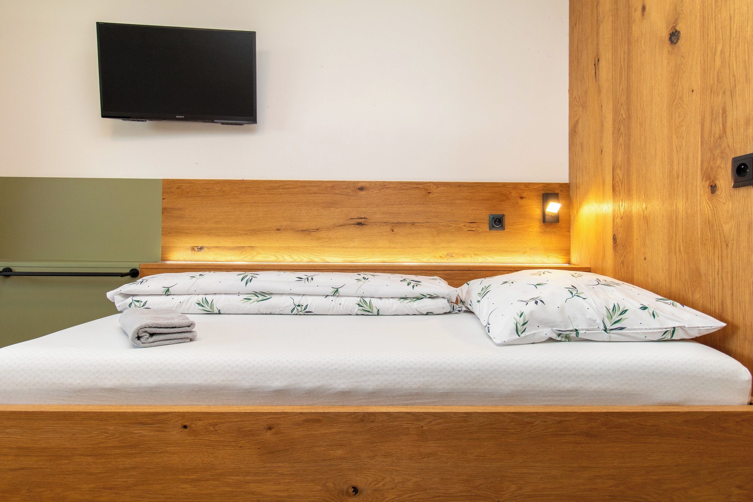 A bed with a wooden headboard, a flat-screen TV mounted on the wall, and a bedside lamp.